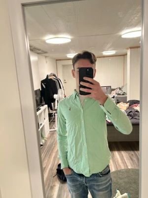 Ljusgrön skjorta från Ralph Lauren - Snygg ljusgrön skjorta från Ralph Lauren med klassisk button-down krage och broderad logga på bröstet. Skjortan har långa ärmar och vita knappar, perfekt för en stilren look.