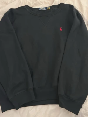 Svart sweatshirt från Polo Ralph Lauren - Klassisk svart sweatshirt från Polo Ralph Lauren med rund halsringning och röd broderad logga på bröstet. Tröjan har ribbade muddar vid ärmslut och nederkant. Perfekt för en avslappnad och stilren look. Storlek 160/100A vilket ungefär motsvarade S. 
