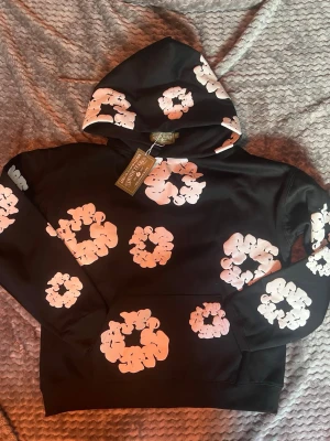 Svart hoodie med vita blommor - Snygg svart hoodie från Market med stora vita blommor i grafiskt mönster över hela tröjan. Hoodien har huva och magficka, perfekt för en trendig streetwear-look. Materialet känns mjukt och skönt, och trycket ger en unik vibe.