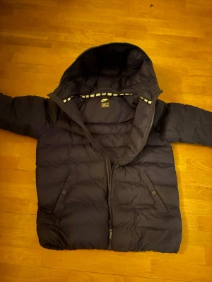 Mörk blå pufferjacka från Nike - Svart pufferjacka från Nike med huva och dragkedja framtill. Jackan har två fickor med knappar och quiltad design som håller värmen. Perfekt för kalla vinterdagar och har en sportig look.