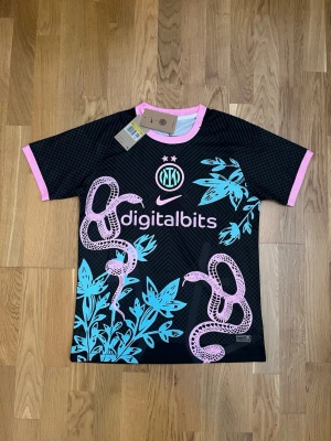 Inter Milan x Nike svart matchtröja - Snygg svart Inter Milan matchtröja från Nike med rosa och blå detaljer. Tröjan har korta ärmar, rosa ormmotiv och blå växtgrafik framtill, samt rosa muddar och krage. Digitalbits-logga på bröstet och klubbmärke. Perfekt för fotboll eller streetwear.