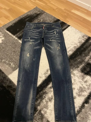 Blå slitna jeans med raka ben - Säljer ett par blå jeans med raka ben och distressed detaljer som slitningar och hål på framsidan. Jeansen har klassisk femficksmodell och en tvättad look med ljusare partier. Perfekta för en avslappnad och trendig stil.