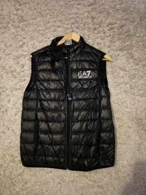 Svart dunväst från EA7 Emporio Armani - Snygg svart dunväst från EA7 Emporio Armani med quiltad design och hög krage. Västen har dragkedja framtill och en tydlig logga på bröstet. Perfekt att slänga över en hoodie eller tröja för en sportig och trendig look. Den är använd 2 gånger, den är som ny. Inga deffekter. 