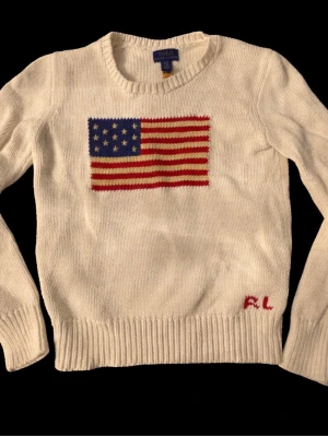 Vit stickad tröja Polo Ralph Lauren - Vit stickad tröja från Polo Ralph Lauren med amerikansk flagga i rött, vitt och blått på bröstet. RL-broderi i rött nedtill. Rund halsringning och ribbstickade muddar vid ärmslut och nederkant. Klassisk och stilren design. Storlek XS