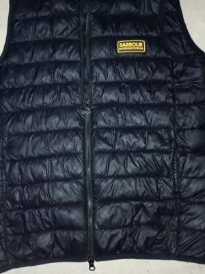 Svart dunväst från Barbour Intl S - Snygg svart dunväst från Barbour International med quiltad design och dragkedja framtill. Västen har en gul logotyp på bröstet och innerficka med knapp. Tillverkad i lätt och värmande material, perfekt för lager-på-lager-stil.