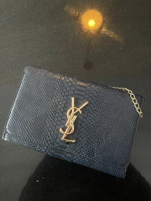Svart kuvertväska från Saint Laurent  - Snygg svart kuvertväska från Saint Laurent i ormmönstrat läder med guldfärgad YSL-logga framtill och en tunn guldkedja. Perfekt accessoar för att lyfta din outfit med en lyxig touch.