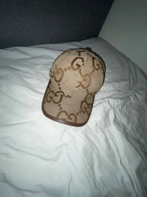 Beige keps från Gucci med GG-mönster - Snygg beige keps från Gucci med det klassiska GG-monogrammet i brunt över hela kepsen. Kepsen har böjd skärm, bruna detaljer och justerbar rem baktill. Materialet är canvas med läderdetaljer för en lyxig känsla.