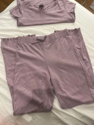 Ljuslila ribbad pyjamas från KappAhl - Mysig tvådelad pyjamas från KappAhl i ljuslila ribbat tyg. Setet består av en kortärmad topp med rund hals och ett par långa byxor med resår och dekorativ rosett i midjan. Båda delarna har vågiga kanter för en söt touch.