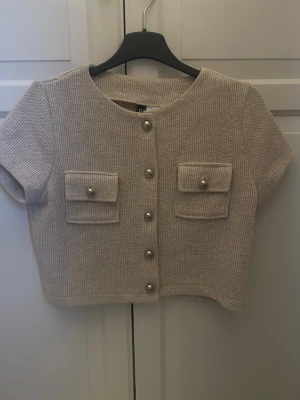 Beige kortärmad kofta från H&M - Trendig beige kortärmad kofta från H&M Divided med rund hals, dekorativa guldfärgade knappar och två fickor framtill. Stickad struktur ger en cool retrovibe och den croppade modellen passar perfekt till höga jeans eller kjol.