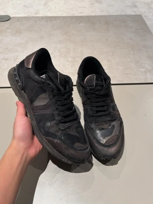 Svarta camo sneakers från Valentino - Snygga svarta sneakers från Valentino med camo-mönster och mattsvarta detaljer. Skorna har lågt skaft, svarta skosnören och en robust sula med Valentino-logga undertill. Materialmix av textil och läder ger en cool och exklusiv vibe.