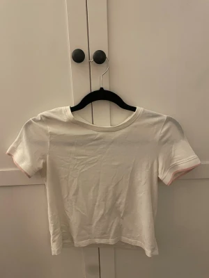 Vit croppad t-shirt med rosa kant - En enkel och stilren vit croppad t-shirt med korta ärmar och diskret rosa kant sopm detalj vid ärmsluten. T-shirten har rund halsringning och är gjord i mjukt bomullstyg. Perfekt till jeans eller kjol för en clean look.🥰💕