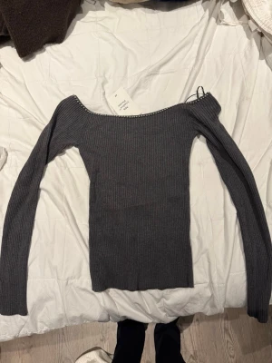 Mörkgrå ribbad offshoulder topp - Trendig mörkgrå ribbad topp med offshoulder-modell och långa ärmar. Toppen har en snygg detalj med små pärlor längs halsringningen och sitter tight mot kroppen. Perfekt för dig som gillar en stilren och modern look.