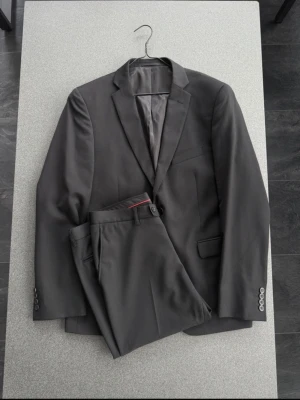 Mörkgrå kostym med tvåknäppt kavaj - Snygg mörkgrå kostym med klassisk tvåknäppt kavaj och matchande byxor. Kavajen har smala slag, tre knappar vid ärmslut och två fickor med lock. Perfekt för dig som vill ha en stilren och tidlös look.