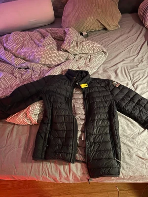 Svart dunjacka från Parajumpers - Snygg svart dunjacka från Parajumpers med gul detalj vid kragen och klassisk patch på ärmen. Jackan har två dragkedjeförsedda fickor och quiltad design som ger en modern look. Perfekt för kalla vinterdagar och riktigt varm. Storlek S