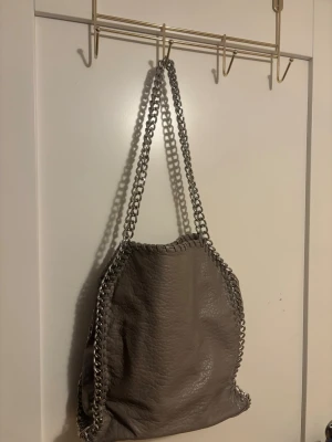 Grå axelväska med kedjedetaljer - Snygg grå axelväska i mjukt, strukturerat konstläder med silverfärgade kedjor längs kanterna och som axelrem. Väskan har en rymlig design och en dragkedjeficka på baksidan. Perfekt för dig som gillar en cool och trendig look.