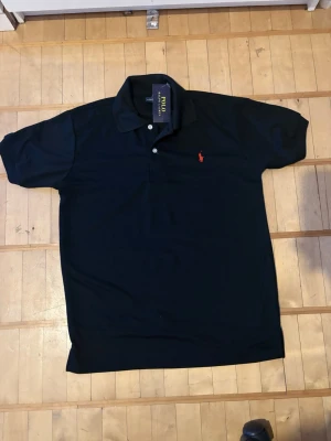 Svart pikétröja från Polo Ralph Lauren - Klassisk svart pikétröja från Polo Ralph Lauren med korta ärmar och krage. Tröjan har två knappar framtill och den ikoniska röda logotypen broderad på bröstet. Perfekt till jeans eller shorts för en clean och stilren look.