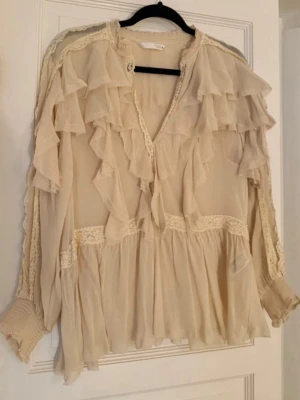 Beige volangblus med spetsdetaljer - Romantisk beige blus med flera lager volanger och spetsdetaljer längs kanterna. Blusen har v-ringning och långa ärmar med breda muddar. Tillverkad i ett tunt, lätt och transparent tyg som ger en luftig känsla.