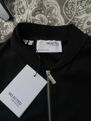 Svart bomberjacka från Selected Homme - Stilren svart full zip tröja från Selected Homme med silverfärgad dragkedja och ribbade muddar. Jackan har en klassisk bomberkrage och två fickor framtill. Perfekt för en clean och modern look.