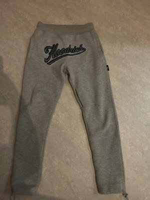 Grå mjukisbyxor från Hoodrich - Säljer ett par grå mjukisbyxor från Hoodrich med stor svart och vit broderad logga framtill. Byxorna har resår i midjan, raka ben och dragsko vid bensluten. Perfekta för chill dagar och streetwear-stil.