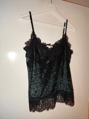 Mörkgrönt sammetslinnemed spets - Snyggt mörkgrönt linne i sammetsmaterial med tunna axelband och dekorativ svart spets vid urringning och nederkant. Perfekt för dig som gillar en lyxig och trendig look. Materialet är mjukt och glansigt, och spetsdetaljerna ger en romantisk touch.