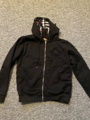 Svart hoodie från Burberry med dragkedja - Säljer en svart hoodie från Burberry med dragkedja framtill och klassiskt rutigt foder i huvan. Hoodien har fickor på magen, justerbar dragsko i huvan och ribbade muddar. Perfekt för dig som vill ha en stilren och exklusiv look.