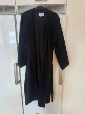 Svart lång kappa med knytskärp - Stilren svart lång kappa med bred krage och knytskärp i midjan. Kappan har en rak passform och trekvartsärmar, perfekt för en trendig och enkel look. Passar till många olika outfits och ger en snygg siluett.