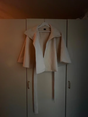 Ljusbeige kappa med bälte - Stilren ljusbeige kappa med bred krage och knytskärp i midjan. Kappan har en öppen design utan synliga knappar och är tillverkad i ett mjukt, tjockt tyg som ger en elegant siluett. Perfekt för dig som gillar minimalistisk och modern stil.