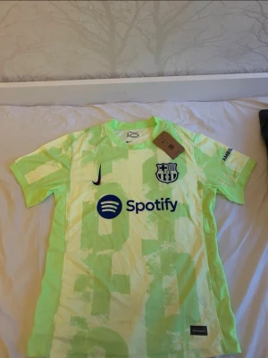 FC Barcelona tränings t-shirt Nike - Snygg FC Barcelona tränings t-shirt i neongult och ljusgrönt med klubbmärke, Nike-logga och Spotify-tryck på bröstet. UNHCR-logga på ryggen. Kortärmad och tillverkad i lätt, ventilerande material. Perfekt för fotbollsträning eller sport.