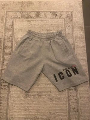 Gråa ICON shorts från Dsquared2 - Snygga grå shorts från Dsquared2 med stor svart 'ICON'-logga och Dsquared2-tryck på ena benet. Mjuk resår i midjan och två sidofickor. Perfekta för en avslappnad och trendig stil.