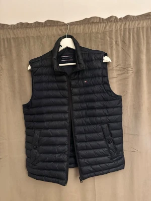 Svart dunväst från Tommy Hilfiger - Snygg svart dunväst från Tommy Hilfiger med quiltad design och hög krage. Västen har dragkedja framtill, två fickor med knappar och en liten logga på bröstet. Perfekt för lager-på-lager och streetstyle.