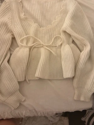 Vit stickad tröja med volang och knyt - Supersöt vit stickad tröja med volangkant längs ringningen och ett knytband i midjan. Tröjan har långa ärmar med breda muddar och en lätt croppad passform. Perfekt för dig som gillar romantisk och trendig stil.