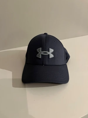 Blå keps från Under Armour - Snygg blå keps från Under Armour med böjd skärm och stort silverfärgat logotryck framtill. Kepsen har även broderad text på sidan och är tillverkad i ett mjukt, sportigt material som andas. Perfekt för dig som gillar stilrena accessoarer.