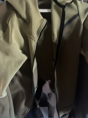 Nike olivgrön hoodie med dragkedja - Snygg olivgrön hoodie från Nike med hel dragkedja och svarta detaljer. Hoodien har en klassisk huva, två sidofickor och Nike-logga på bröstet. Tillverkad i mjukt och bekvämt material, perfekt för en avslappnad streetwear-look.