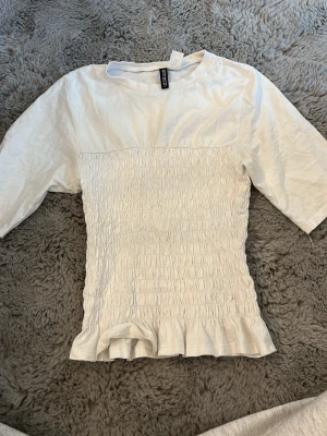 Vit smockad topp från H&M Divided - Supersöt vit topp från H&M Divided med smockad midja och volangkant nedtill. Toppen har korta ärmar och är gjord i mjuk bomull. Perfekt att styla med jeans eller kjol för en trendig look.
