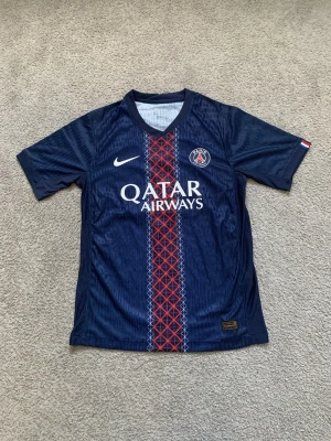 PSG matchtröja Nike Dembélé #10 - Mörkblå PSG matchtröja från Nike med O.Dembélé och nummer 10 på ryggen. Tröjan har Qatar Airways-tryck på bröstet, klubbmärke och vit Nike-logga. Röd och vit detalj i mitten samt korta ärmar. Tillverkad i lätt och ventilerande material. Priset är ej hugget i sten och kom gärna med frågor!