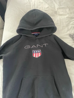 Svart GANT hoddie  - Säljer en svart hoodie från GANT med stor broderad logga och sköld med stjärnor och ränder på bröstet. Klassisk känguruficka framtill och huva. Perfekt för dig som gillar stilrena och sportiga plagg. Materialet är mjukt och bekvämt, troligen bomullsblandning.
