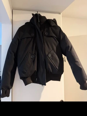 Svart Nobis vinterjacka med huva Dam  - Snygg svart bomberjacka med stor huva och breda muddar. Jackan har fyra stora fickor framtill med dekorativa knappar och dragkedja. Perfekt för dig som vill ha en stilren och funktionell jacka. Damjacka storlek xl men funkar till både tjej och Kille 