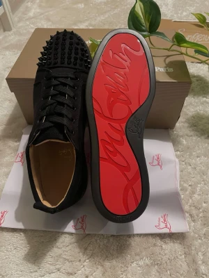 Svarta Louboutin skor  - Snygga svarta sneakers från Christian Louboutin med ikonisk röd sula och svarta nitar på tån. Skorna har snörning och är tillverkade i läder med textildetaljer. Perfekta för dig som vill sticka ut med en lyxig och edgy stil.