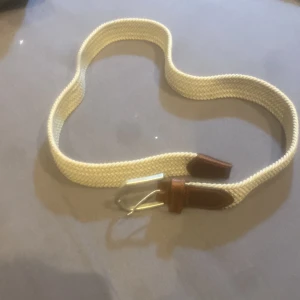 Beige flätat bälte med läderdetaljer - Snyggt beige flätat bälte med bruna läderdetaljer och silverfärgat spänne. Perfekt accessoar för att lyfta din outfit och ge en stilren touch. Passar till både jeans och chinos.