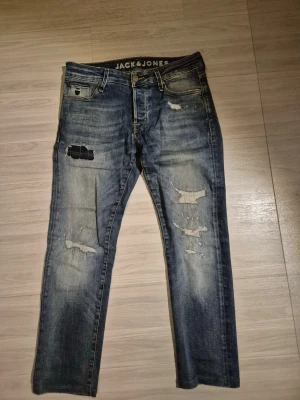 Blå slitna jeans från Jack & Jones - Säljer ett par blå jeans från Jack & Jones med slitningar och trasiga detaljer på benen. Jeansen har klassisk femficksmodell, normal midja och raka ben. Materialet är denim i bomull med en snygg tvättad look och patch bak i brunt läder. Storleken är W30 L30, men skulle säga att den passar w30 28L. Passformen är som slim fit 