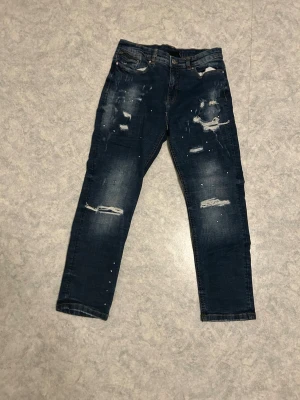 Mörkblå slitna jeans med hål - Snygga mörkblå jeans med slitningar och hål på framsidan. Jeansen har en klassisk femficksmodell och raka ben. Perfekta för en trendig och avslappnad look. Materialet är denim och färgen är tvättad mörkblå med ljusa detaljer.