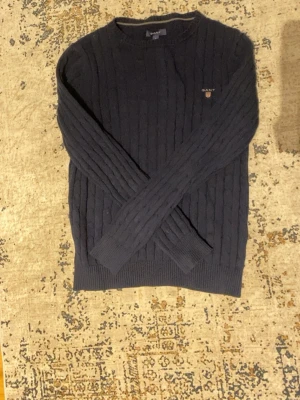 Mörkblå kabelstickad tröja GANT - Kabelstickad mörkblå tröja från GANT med rund halsringning och broderad logotyp på bröstet. Tröjan har långa ärmar och ribbade muddar vid ärmslut och nederkant. Perfekt för dig som gillar klassisk och stilren design.