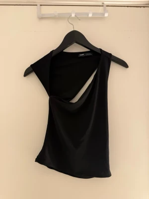 Svart asymmetrisk topp  - Säljer en svart, glansig och asymmetrisk topp. Toppen har en unik draperad design med en axel bar och en cool snedskuren halsringning. Materialet är mjukt och stretchigt, perfekt för att sticka ut med en trendig look.