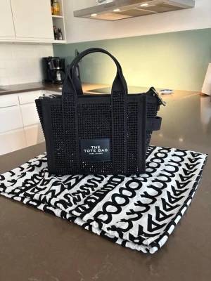 Svart The Tote Bag från Marc Jacobs - Äkta från raglady. Snygg svart handväska från Marc Jacobs, modell The Tote Bag. Väskan är dekorerad med svarta nitar över hela ytan och har både handtag och axelrem. Märkeslogga framtill och levereras med en mönstrad dustbag. Perfekt för dig som gillar statement-accessoarer.