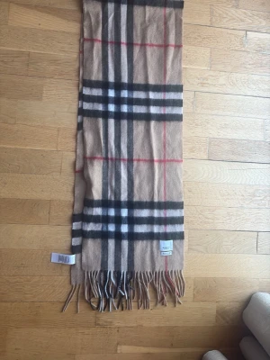 Beige rutig Burberry halsduk kashmir - Klassisk beige halsduk från Burberry i 100% kashmir. Den har det ikoniska rutiga mönstret i svart, vitt och rött samt fransar i ändarna. Perfekt accessoar för att lyfta din outfit med en tidlös brittisk vibe.