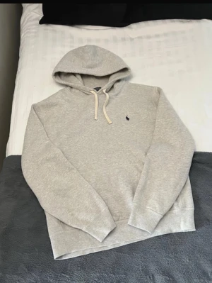 Grå hoodie från Polo Ralph Lauren - Snygg ljusgrå hoodie från Polo Ralph Lauren med klassisk huva och vita dragsnören. Diskret broderad logga i marinblått på bröstet. Tillverkad i mjuk bomullsblandning som känns riktigt skön mot huden. Perfekt för en avslappnad och stilren look.