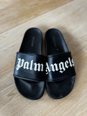 Svarta Palm Angels sandaler - Svarta slides från Palm Angels med bred rem över foten och vit logotyp i gotisk stil. Tillverkade i syntetmaterial med bekväm innersula och klassisk slip-on design. Perfekta för en avslappnad och trendig look.