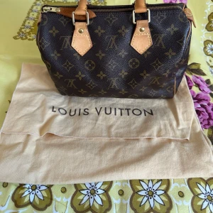 Louis Vuitton Speedy Monogram handväska - Säljer en klassisk Louis Vuitton Speedy handväska i brunt monogram canvas med ljusbruna läderhandtag och guldfärgade metalldetaljer. Väskan har dragkedja upptill och ett rymligt innerfack med brunt tygfoder. Perfekt för dig som gillar ikoniska designeraccessoarer.