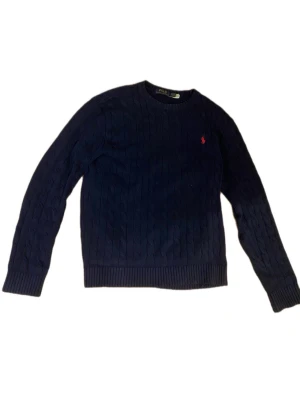 Mörkblå kabelstickad tröja Polo Ralph Lauren - Snygg mörkblå kabelstickad tröja från Polo Ralph Lauren med rund halsringning och röd broderad logga på bröstet. Tröjan har långa ärmar och ribbade muddar vid ärmslut och nederkant. Perfekt för en stilren och klassisk look.