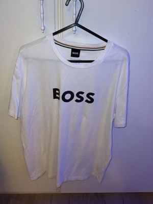 Vit BOSS t-shirt med tryck - Vit t-shirt från BOSS med svart logotyp-tryck på bröstet. Klassisk rund halsringning och korta ärmar. Tillverkad i mjuk bomull för en skön och avslappnad känsla. Perfekt för en stilren och enkel look.